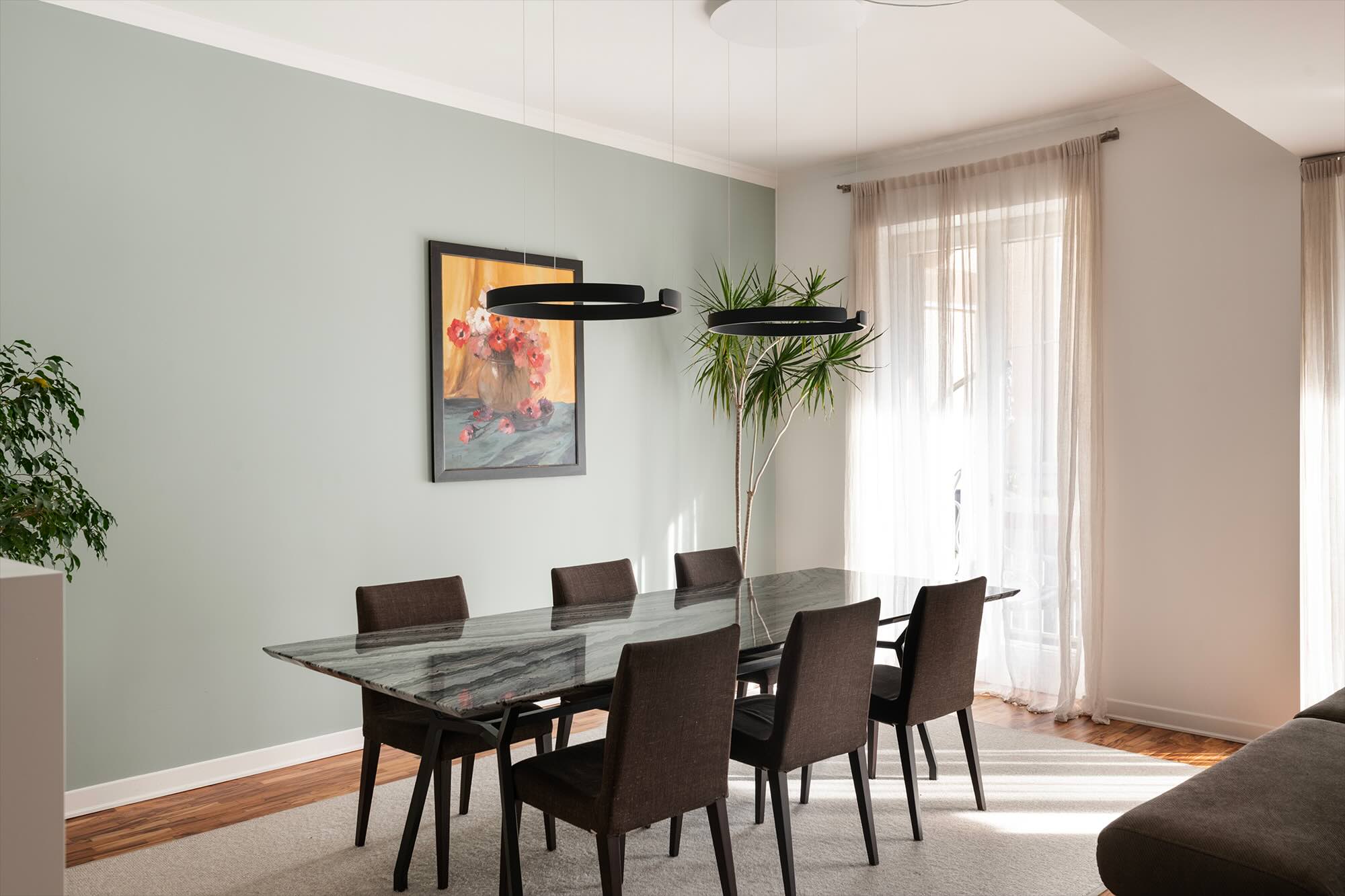 ristrutturazione di una casa a Bergamo con tavolo in marmo, sedie in tessuto scuro e pareti in verde salvia, illuminata da luce naturale e lampade sospese di design