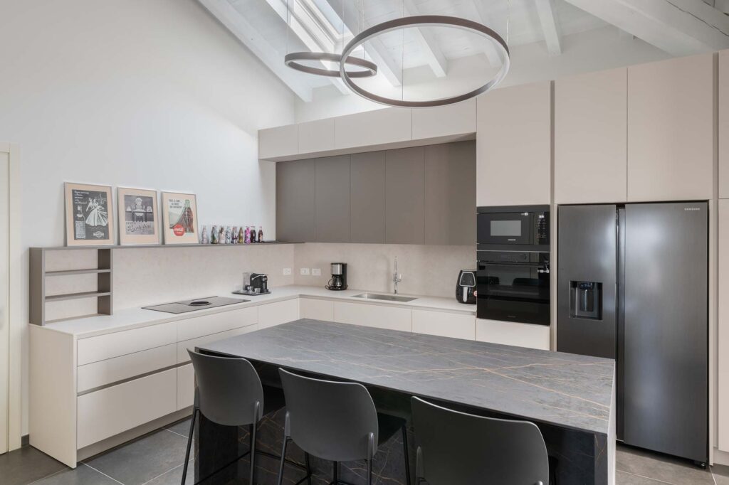 Ristrutturazione cucina a Bergamo con isola in marmo scuro, arredi su misura beige, elettrodomestici a incasso e illuminazione moderna a sospensione.